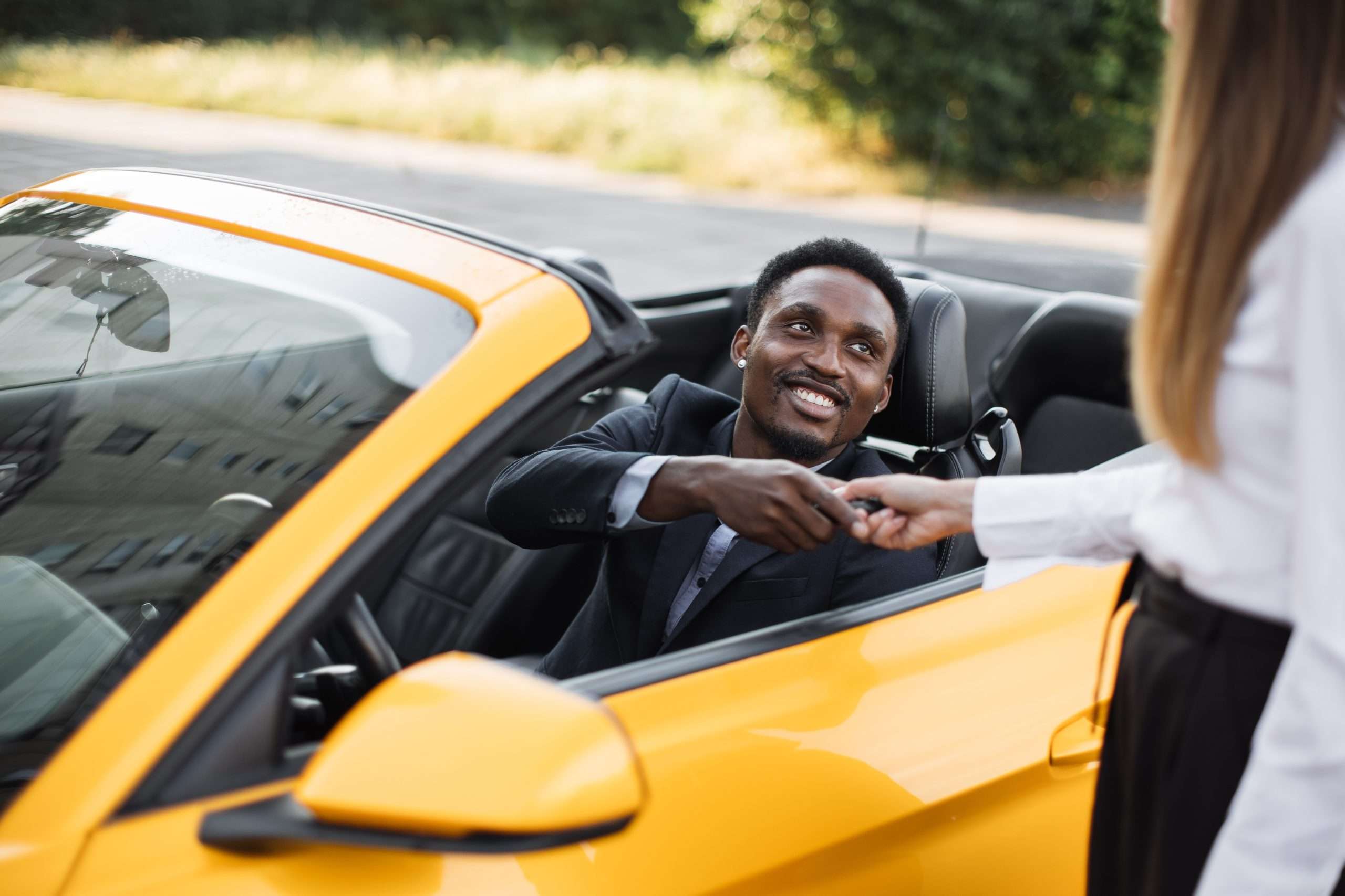 A black man in a yellow sportscar shaking hands with a woman in the foreground
